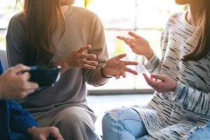 Three individuals engaged in a supportive group conversation, with one person holding a coffee cup, representing a peer support program setting.