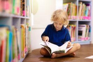 Young child sitting on the floor in a library, reading a book surrounded by bookshelves - illustrating speech development activities for children through reading.